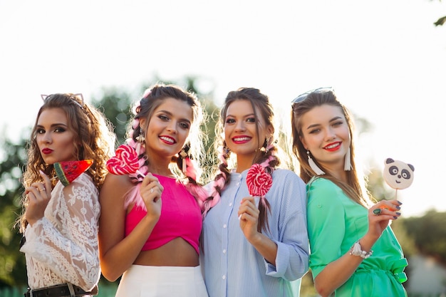 Group of pretty and fashionable girls wearing in trendy clothes with colorful braid and professional makeup, walking at street after shopping. Women holding candies heart on stick and smiling.