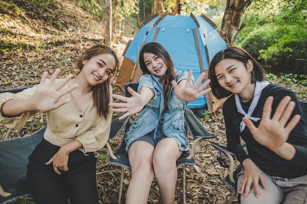 Group of pretty women sitting on camping chair at front of tent
for relaxtion they waving hands and looking camera to photography
while camping in nature forest with happiness together copy
space