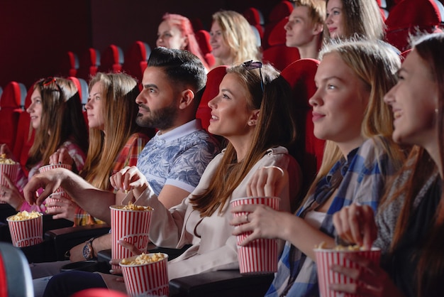 Group of pretty and young female watching new entertaining comedy in cinema hall. Cheerful young blonde laughing, eating popcorn and enjoying free time on weekend. Concept of happiness and fun.