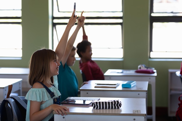 Group of school children sitting at desks and raising their hands to answer