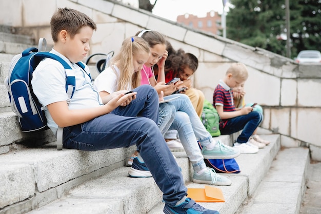 Group of schoolchildren in colorful clothes are sitting on the steps and looking at gadgets. Seven classmates of different nationalities.