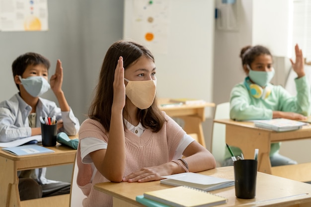 Group of schoolchildren in masks raising hands at lesson