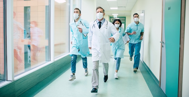 Group of serious and confident doctors in safety medical masks are running fast on the hospital for an urgent sick patient