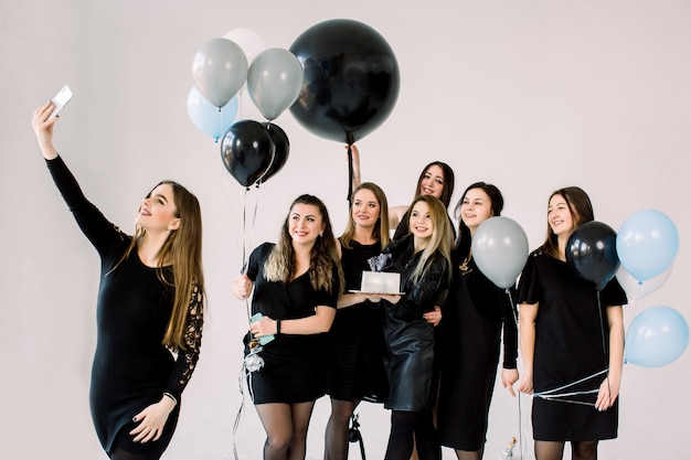 Group of seven girls best friends making selfie on smartphone, enjoying birthday party with cake and air balloons, having fun, smiling and laughing. Friendship and birthday party concept