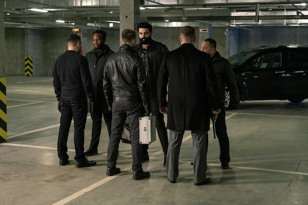 Group of six intercultrual men in black standing in front of each other on parking area while discussing terms of criminal deal