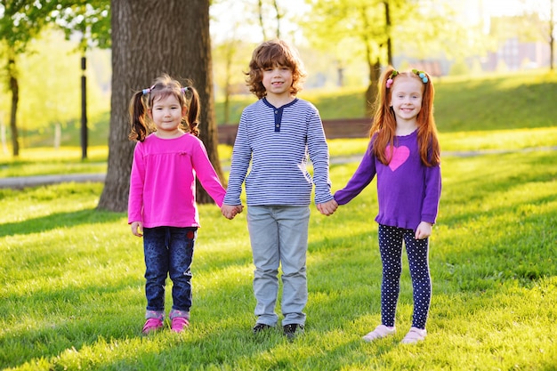 A group of small children smiling holding hands on a of grass, a tree and a park.