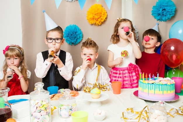 Группа улыбающихся детей, играющих на дне рождения в украшенной комнате. Счастливые дети дуют в трубы на дне рождения
