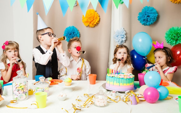 Group of smiling children playing on the birthday party in decorated room. Happy kids blowing in pipes on birthday party