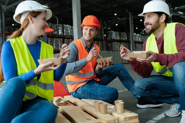 Group smiling workers wearing hard hats vests holding lunch boxes eating lunch sitting talking