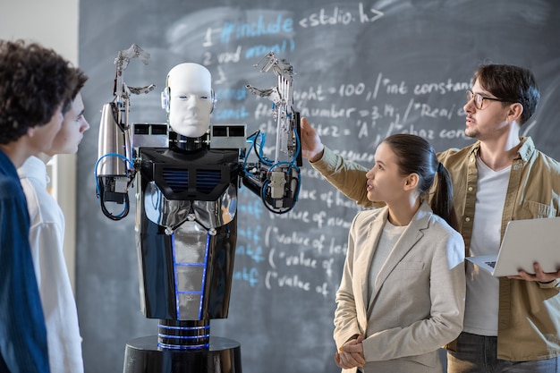 Group of students looking at computer control robot with raised hands while their teacher demonstrating its abilities at lesson