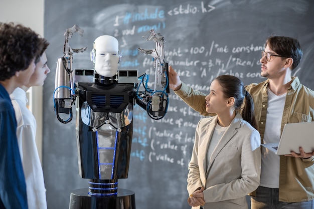 Photo group of students looking at computer control robot with raised hands while their teacher demonstrating its abilities at lesson