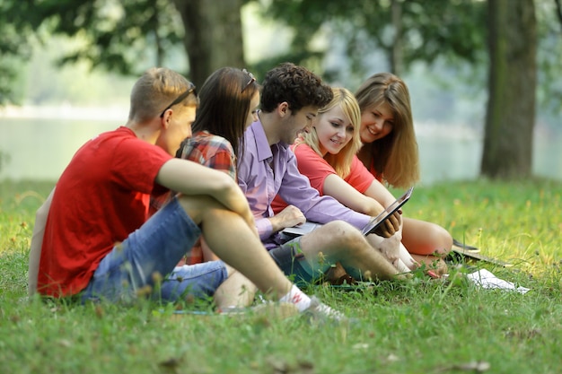 Photo group of students with a laptop sitting on the grass in the park.