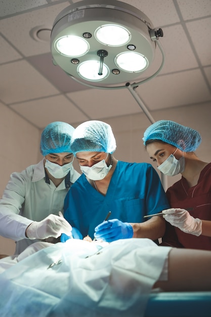 Группа хирургов за работой в хирургическом театре. Команда медицины реанимации нося защитные маски держа стальные медицинские инструменты сохраняя пациента. Хирургия и неотложная помощь.