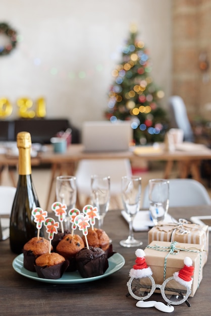 Group of tasty homemade muffins with small xmas cards on plate surrounded by bottle of champagne, Santa eyeglasses, flutes and giftboxes