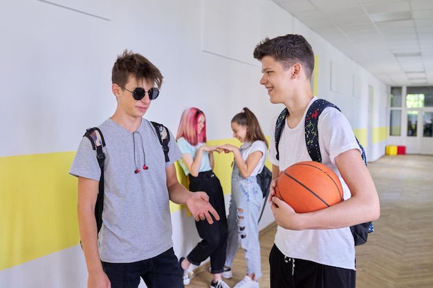 Group of teenage students talking standing outside school building two males with backpacks basketball ball in focus schoolgirls in corridor background Lifestyle communication youth concept