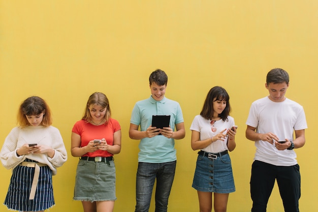 Group of teenagers using their mobile phones and tablet on a yellow background.