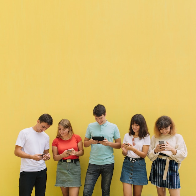 Group of teenagers using their mobile phones and tablet on a yellow background.