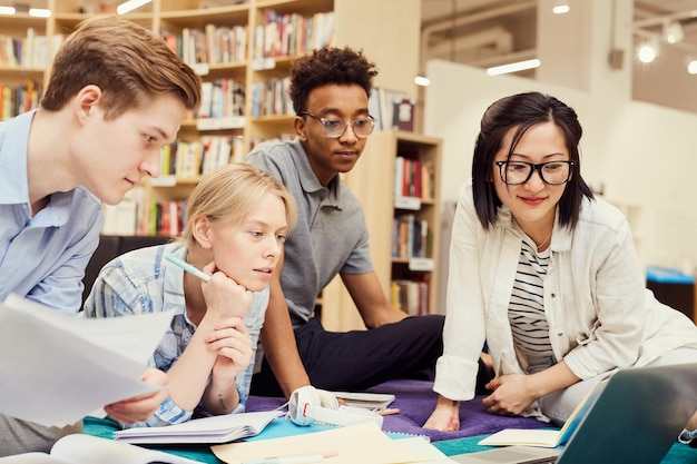 Photo group of thoughtful young multiethnic students focused on joint project sitting among workbooks and devices in library and looking at laptop screen