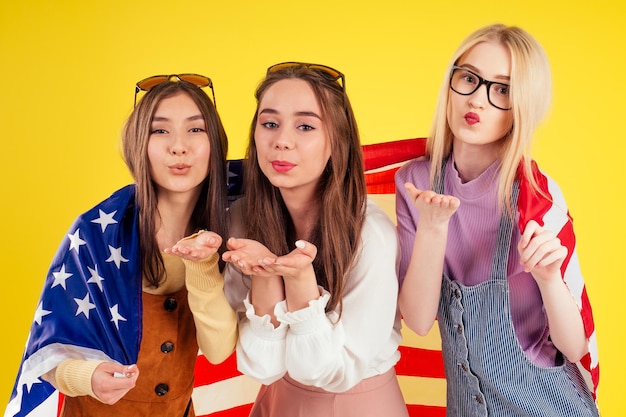 Group of three Asian and European laughing girls holding passports and tickets photo camera wrapped in american flag spring party summer style yellow background studio,visa and independence day usa