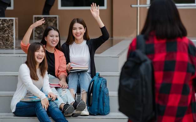 세 명의 매력적인 아시아 대학생 그룹이 대학 캠퍼스의 계단에 앉아 야외에서 친구들에게 인사를 건네고 있습니다. 교육, 우정, 대학생들의 삶에 대한 개념.