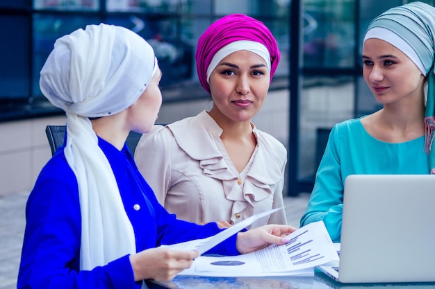 Group of three caucasian Muslim office lady discussing with a businesswoman partner job interview shawl and turban on head perfect skin and makeup sitting at a round table with a laptop and papers