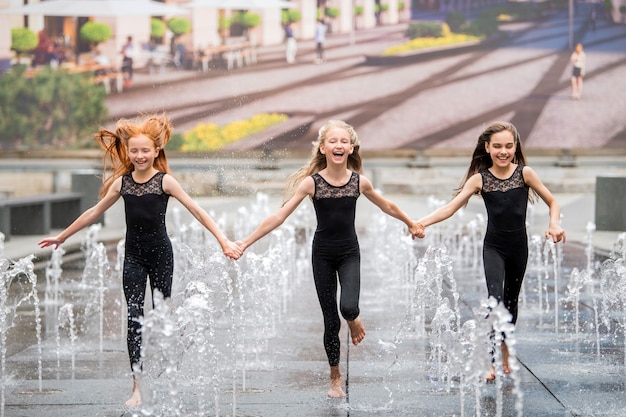 A group of three little ballerinas in black tight-fitting suits run to the viewer amid splashing fountains against backdrop of a cityscape on hot day.