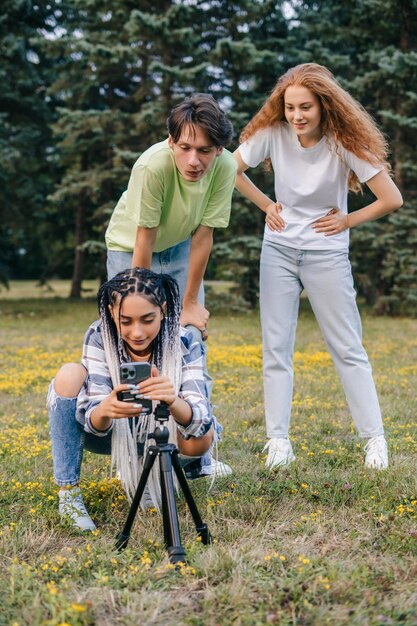 Photo group of three young teenagers friends wanting to take a group selfie with a selfie stick standing in city park social media trends friendship day