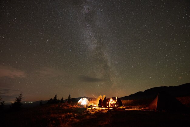 Group of tourists with guitar by burning bonfire under dark starry sky with Milky Way constellation.