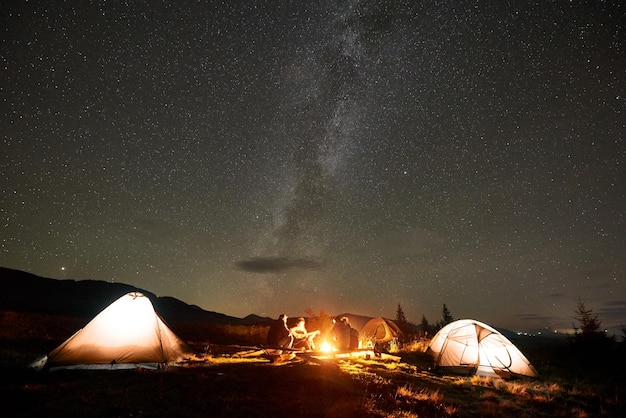 Group of tourists with guitar by burning bonfire under dark starry sky with Milky Way constellation.