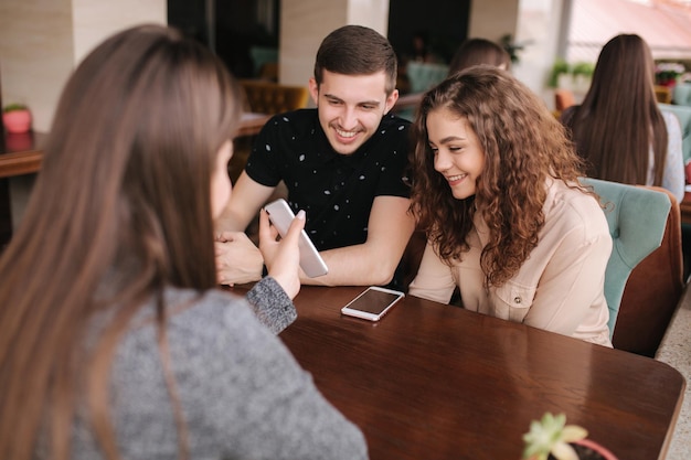 Group of tree friends on terrace Yound people meeting in cafe Friends sitting in cafe together Female show to couple something on her smartphone