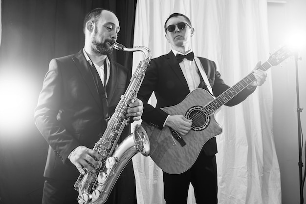 Group of two musicians, male jazz band, guitarist and\
saxophonist in classical costumes improvise on musical instruments\
in a studio black and white shot