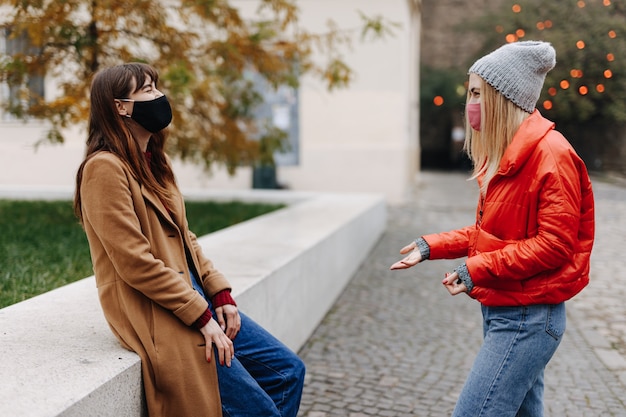 Group of two young women dressed in casual warm clothing talking on smiling on street. Happy friends wearing protective face masks during pandemic time.