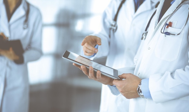 Group of unknown doctors use a computer tablet to check up some medical names records, while standing in a hospital office. Physicians ready to examine and help patients. Medical help, insurance in he