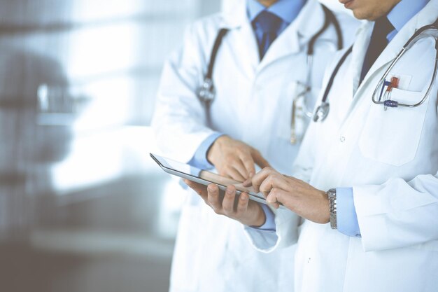 Group of unknown doctors use a computer tablet to check up some medical names records, while standing in a hospital office. Physicians ready to examine and help patients. Medical help, insurance in he