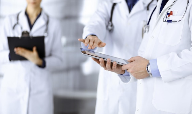 Group of unknown doctors use a computer tablet to check up some\
medical names records, while standing in a hospital office.\
physicians ready to examine and help patients. medical help,\
insurance in he