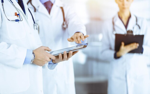 Group of unknown doctors use a computer tablet to check up some medical names records, while standing in a sunny hospital office. Physicians ready to examine and help patients. Medical help, insurance