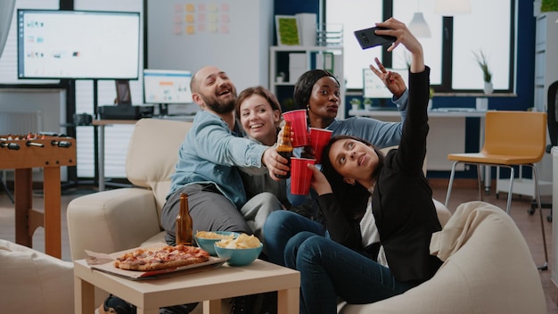 Group of workmates clinking glasses of beer after work and using smartphone to take selfie at office party. Colleagues enjoying fun activity with snacks and drinks for entertainment.