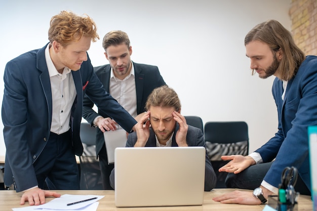 Group of young adult men in suits gathered together near laptop working in office