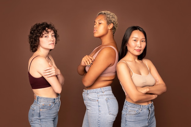 Group of young beautiful females in tanktops and blue jeans standing in row against brown background in front of camera and looking at you