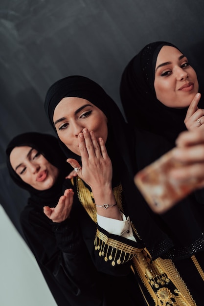 Group of young beautiful muslim women in fashionable dress with hijab using smartphone while taking selfie picture in front of black background.