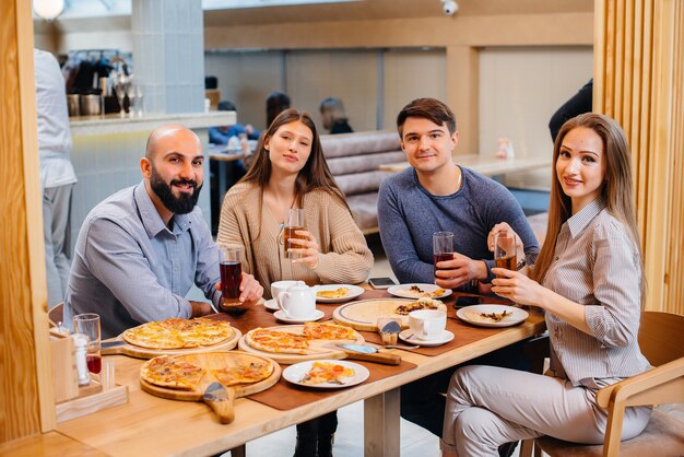 A group of young cheerful friends is sitting in a cafe talking and eating pizza. Lunch at the pizzeria.