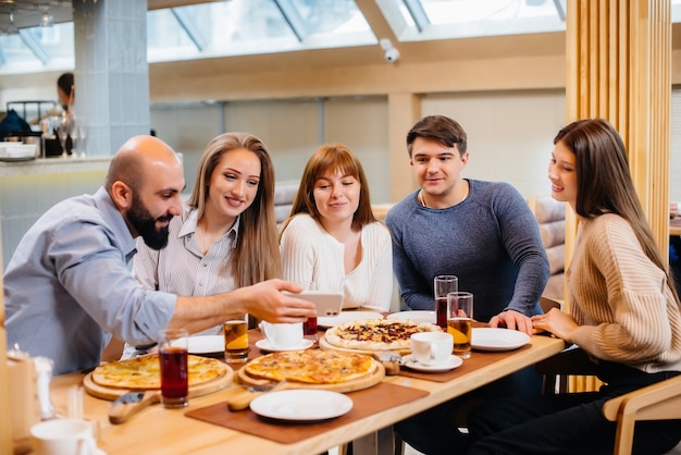 Группа молодых веселых друзей сидит в кафе, разговаривает и ест пиццу