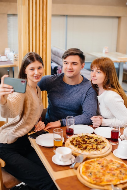 A group of young cheerful friends is sitting in a cafe talking and taking selfies on the phone. Lunch at the pizzeria.