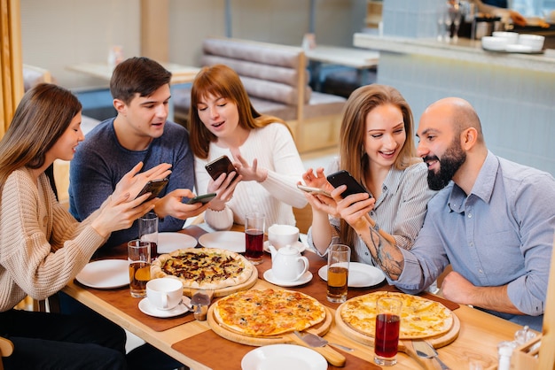 A group of young cheerful friends is sitting in a cafe talking and taking selfies on the phone. Lunch at the pizzeria.