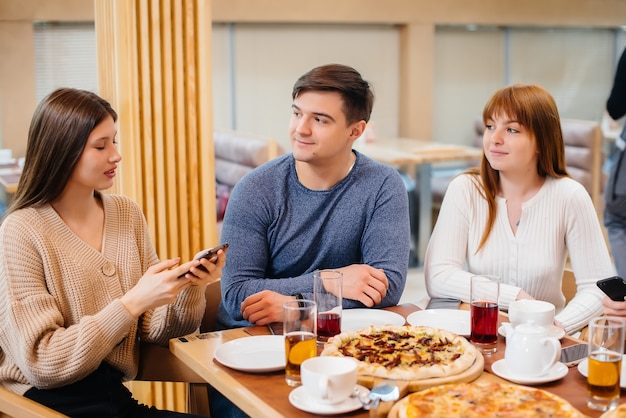 A group of young cheerful friends is sitting in a cafe talking and taking selfies on the phone. Lunch at the pizzeria.