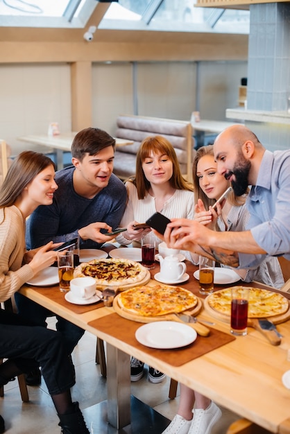 A group of young cheerful friends is sitting in a cafe talking and taking selfies on the phone