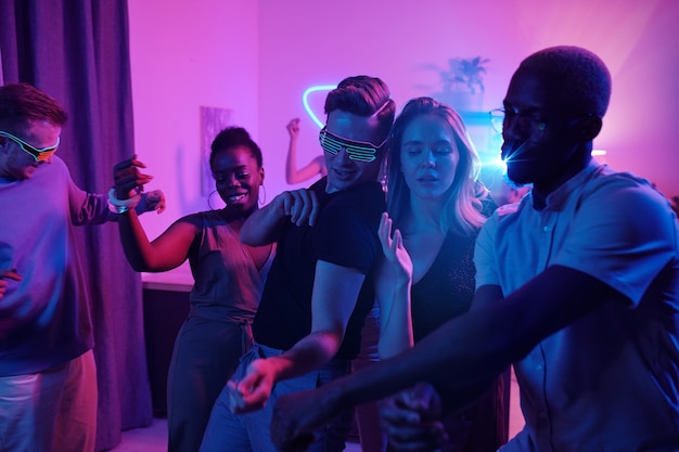 Group of young cheerful intercultural friends in smart casualwear dancing excitedly at home party in living-room illuminated with pink lighting