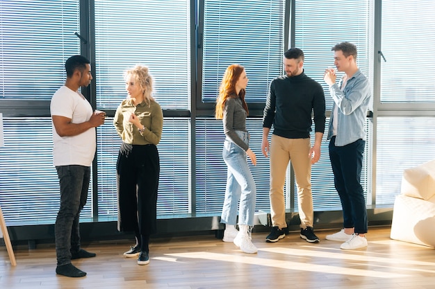 Group of young creative multiethnic coworkers spending time together in modern office chatting and discussing upcoming projects. Front view of diverse colleagues group talking at work break.