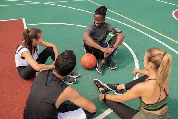 Photo group of young friendly intercultural people sitting on playground while getting ready for another game of basketball