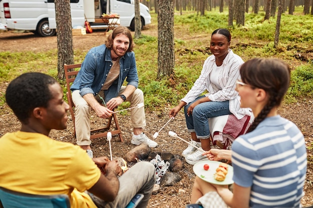 Un gruppo di giovani amici che friggono marshmallow sul fuoco all'aperto durante il campeggio si siedono intorno al ca